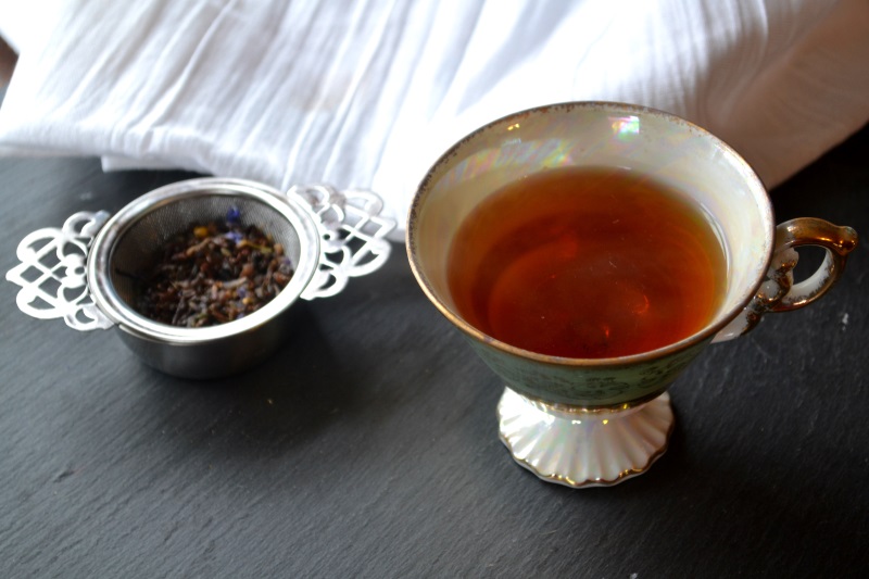 Cup of Blue Moon tea sitting on gray surface with strainer of tea leaves next to white cloth. from www.ApronFreeCooking.com