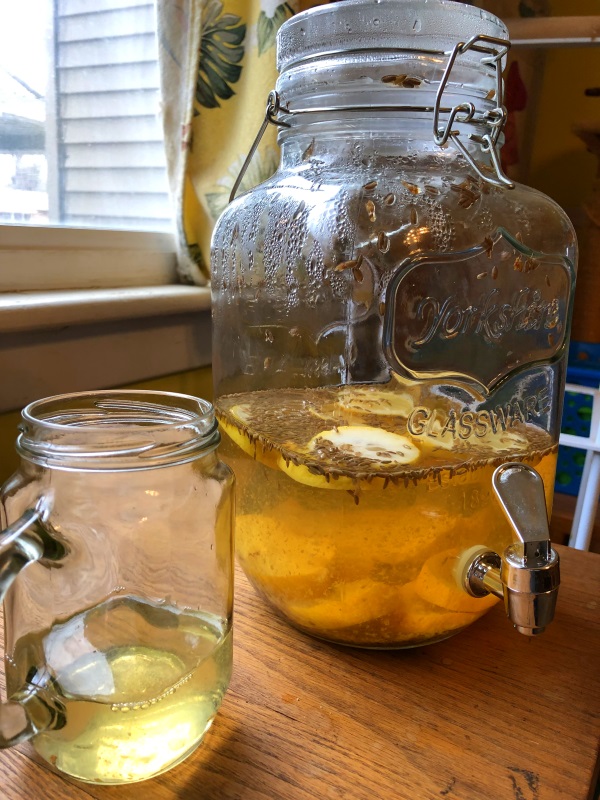 Glass of lavender lemonade in front of large jar containing lavender lemonade from www.ApronFreeCooking.com