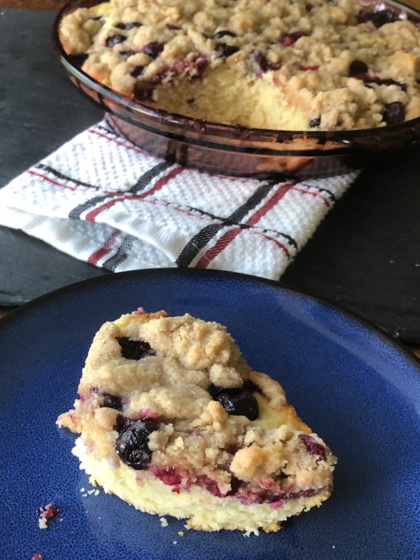 Old Fashioned Blueberry Buckle with crumb topping. Single serving on a blue plate in front of purple pie plate full of blueberry buckle coffee cake. 